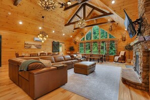 A log cabin interior with a large L-shaped sectional sofa, a stone fireplace, and a dining area with a view of the forest.