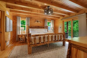 Een slaapkamer in een houten blokhut met een groot bed, een houten hoofdeinde, een plafondventilator en uitzicht op groen.