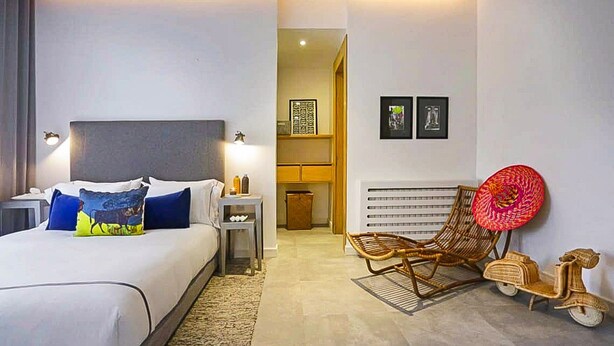 Una habitación de hotel moderna con cama, mesitas de noche, una silla, una pequeña mesa y un radiador fijado a la pared.