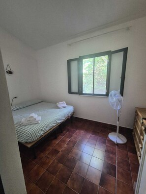 A small, single-room accommodation with a bed, a window, and a standing fan. - A Wonderful Villa Surrounded by Lush Greenery, Peacefully Located Near the Beach (Calabria)