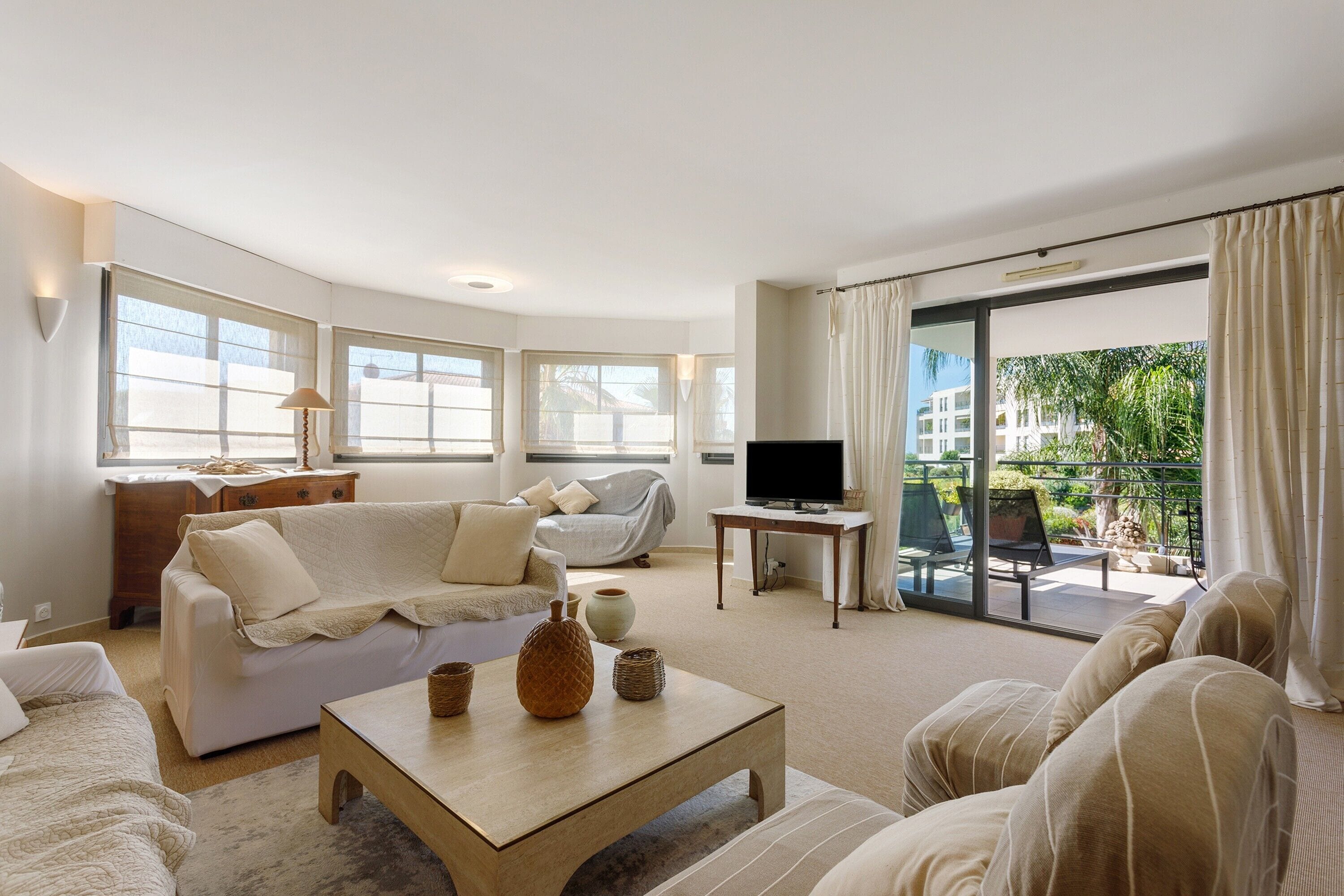 A modern living room with a sofa, a coffee table, and a television. There is a balcony with a view of greenery.