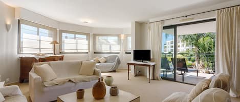 A modern living room with a sofa, a coffee table, and a television. There is a balcony with a view of greenery.