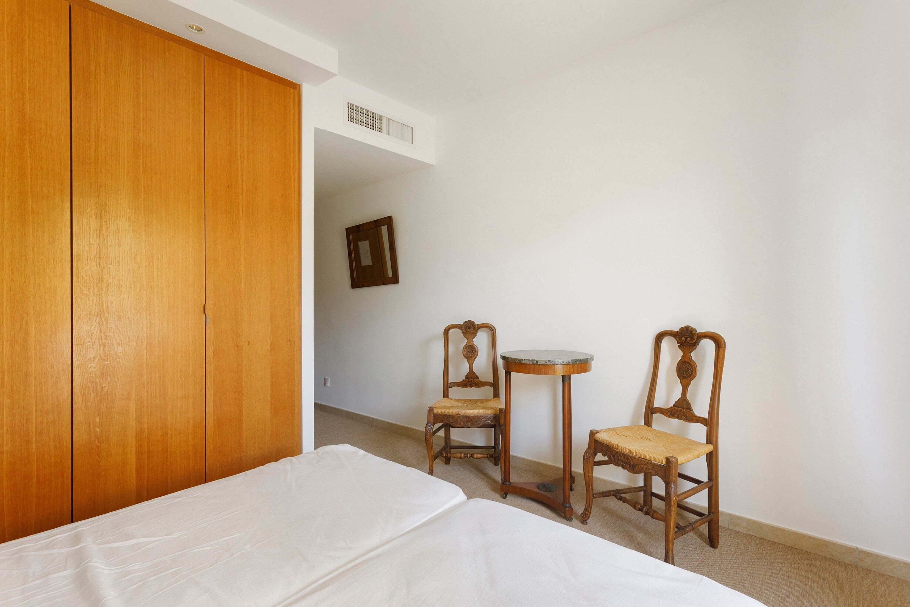 A hotel room with a wooden wardrobe, a bed with white bedding, two wooden chairs, a small round table, and a wall-mounted mirror.