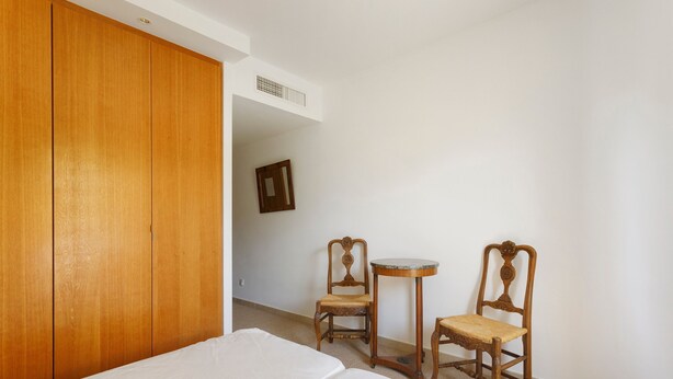 Habitación de hotel con un armario de madera, una cama con ropa de cama blanca, dos sillas de madera, una mesita redonda y un espejo montado en la pared.