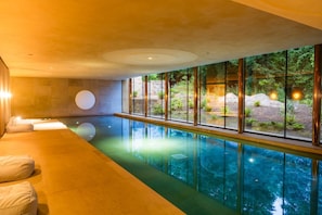 Une piscine intérieure moderne offrant une vue dégagée sur l’extérieur, un espace détente avec des coussins et un mur en pierre.