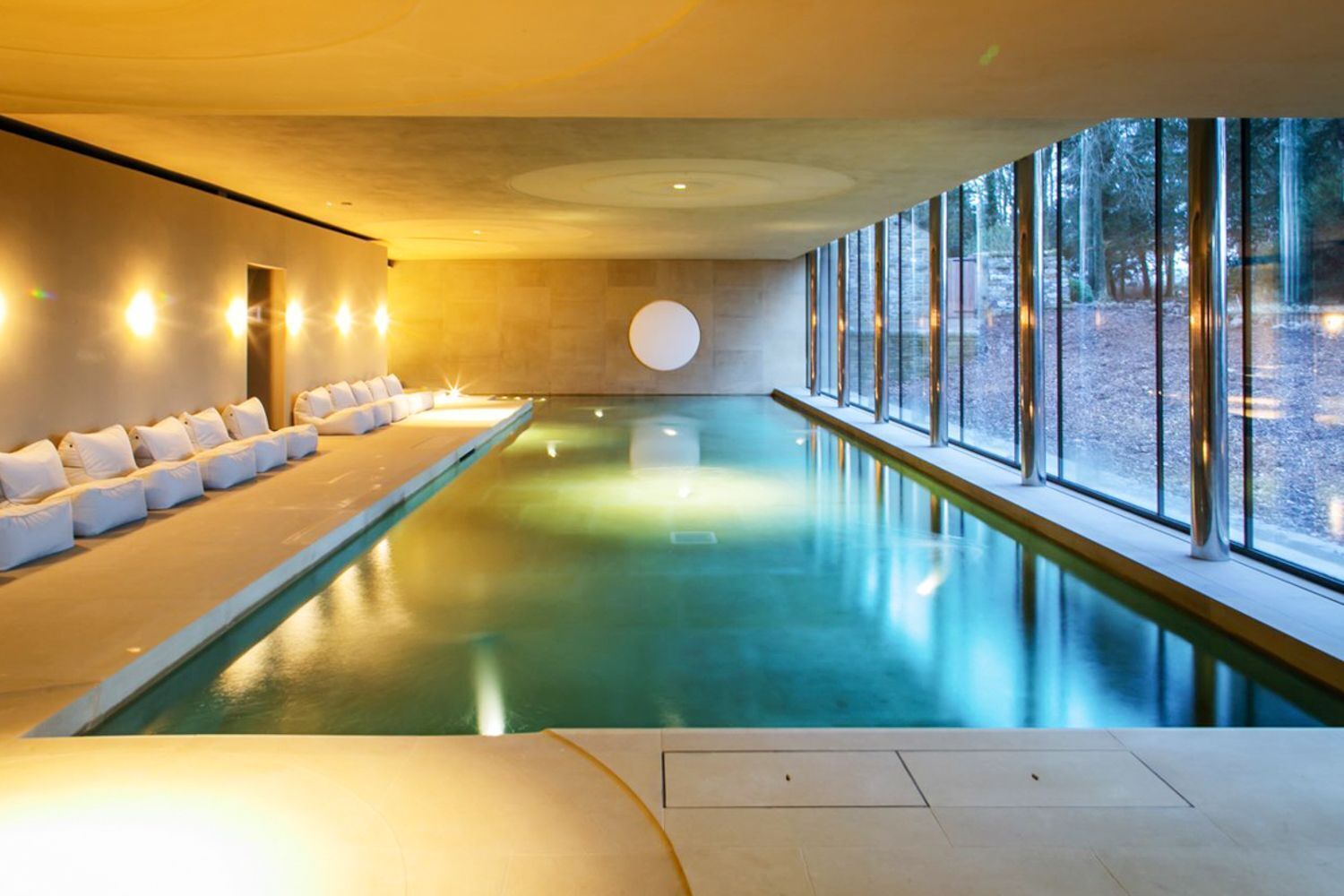 A modern indoor swimming pool with a sunlit interior, a long seating area with cushions, and large windows offering a view of the outdoors.