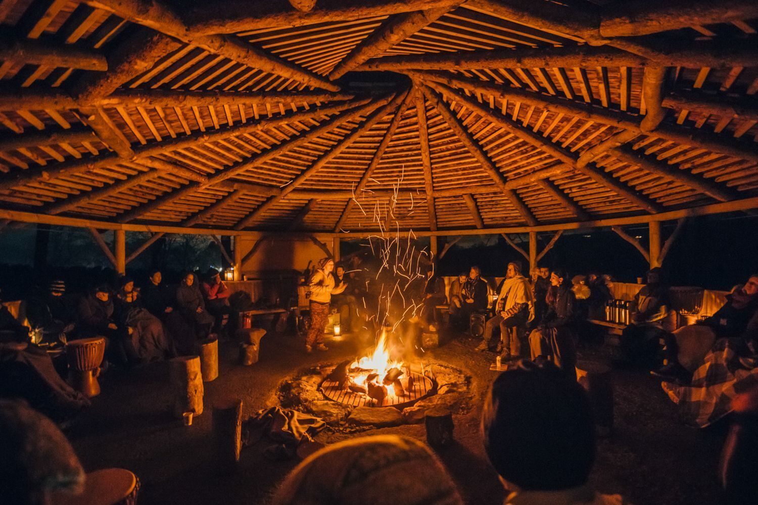 Eine gesellige Runde im Freien um ein Feuer, überdacht mit einer Holzkonstruktion.