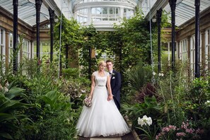 Una pareja en un invernadero con techo de cristal, rodeada de abundante vegetación y flores.