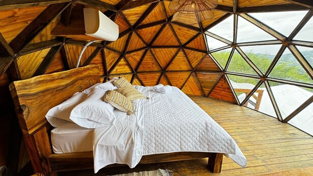 Una habitación con techo y paredes de madera, cama con ropa blanca y vistas amplias a través de grandes ventanales.