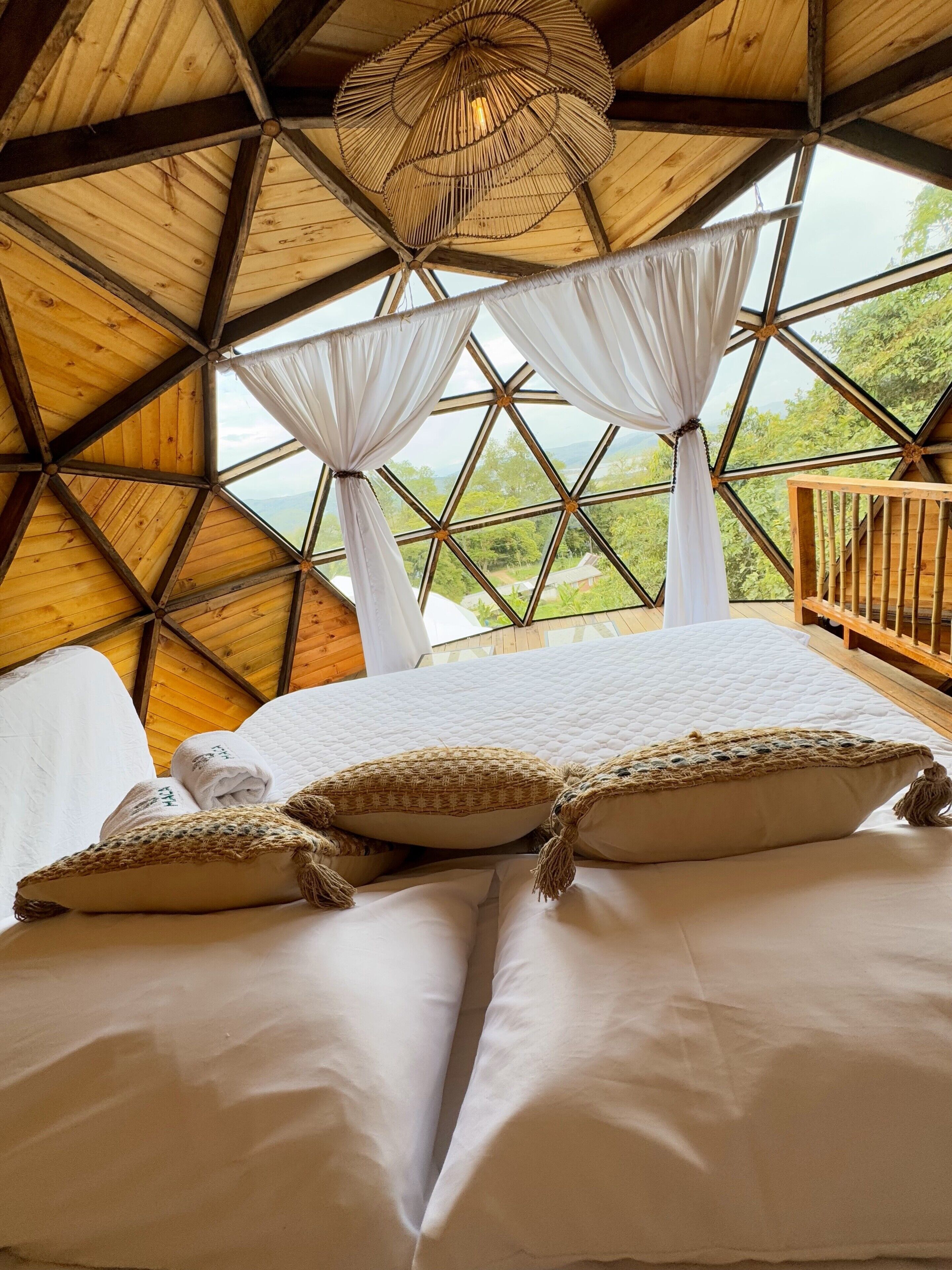 A room with a large bed, white curtains, and a view of greenery through a geometric window.
