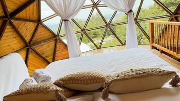 A room with a large bed, white curtains, and a view of greenery through a geometric window.