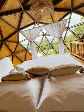A room with a large bed, white curtains, and a view of greenery through a geometric window.