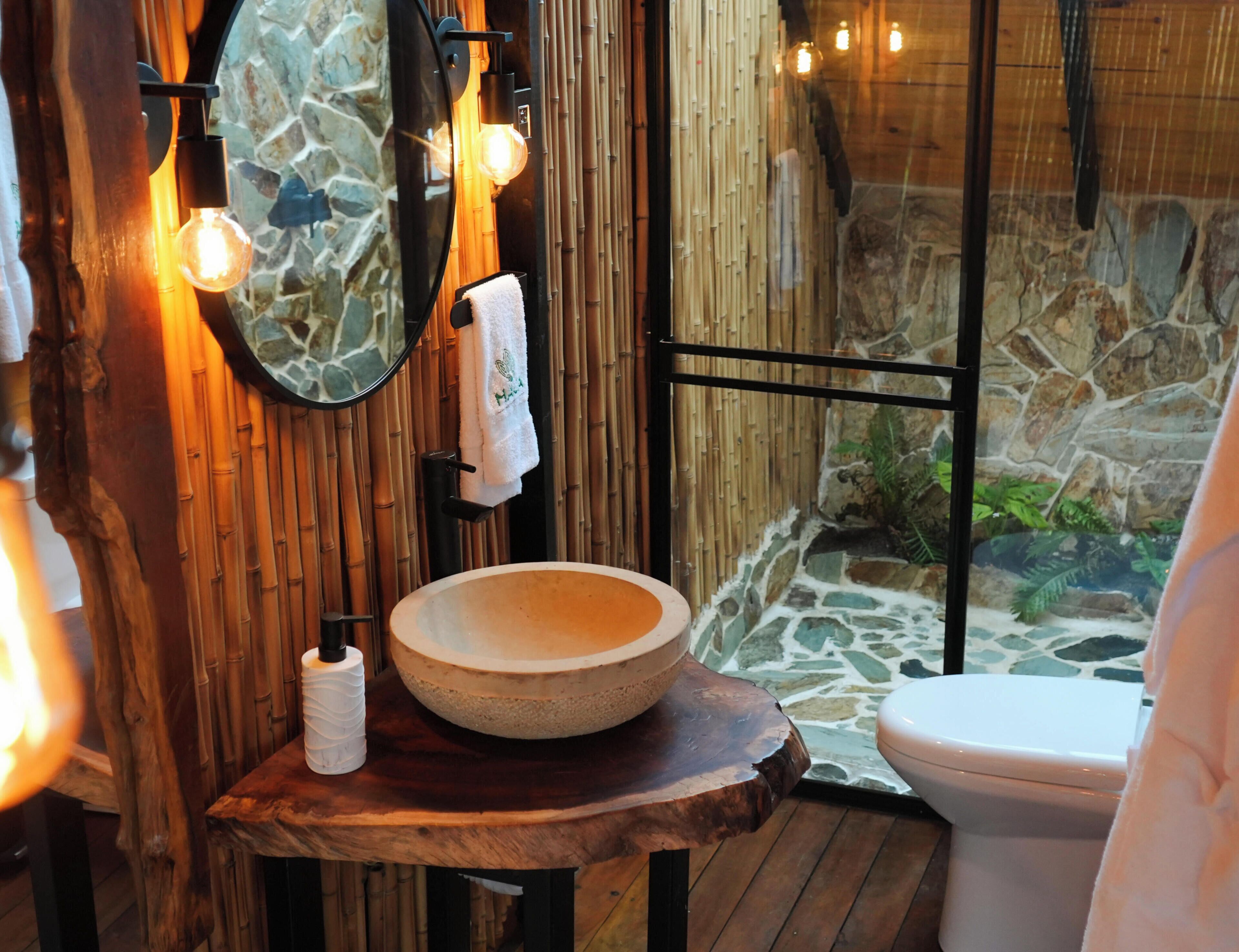 A bathroom with a stone wall, a wooden vanity, a round basin, a toilet, and a glass shower enclosure.