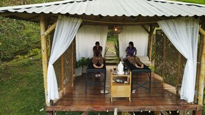 A covered outdoor relaxation area with two massage tables, a small table with a bottle and glass, and a decorative light fixture. - MACA glamping (Calima)