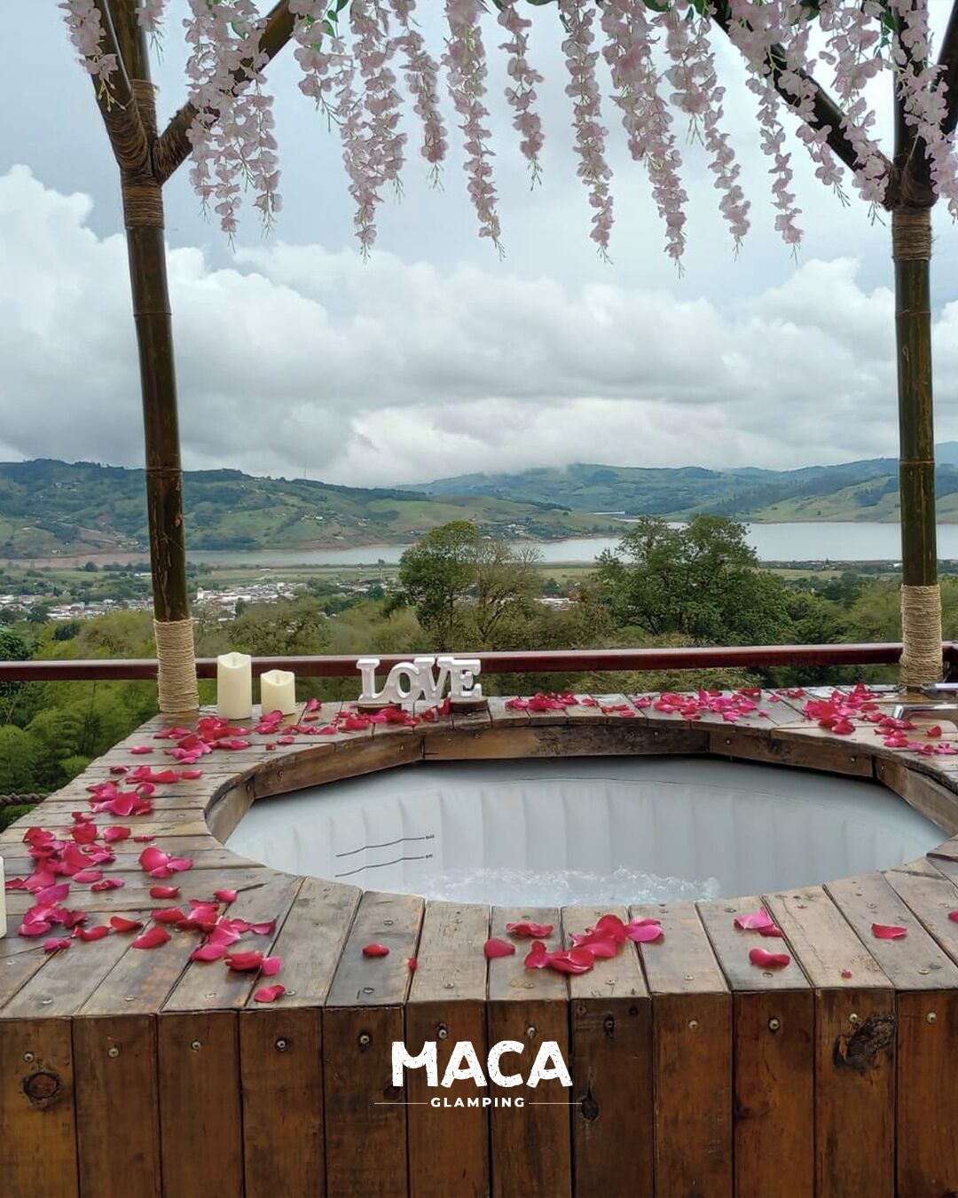 Tinaco con cubierta de madera y pétalos rosados esparcidos, rodeado por una pérgola de madera con flores rosadas, con vistas a un paisaje pintoresco.