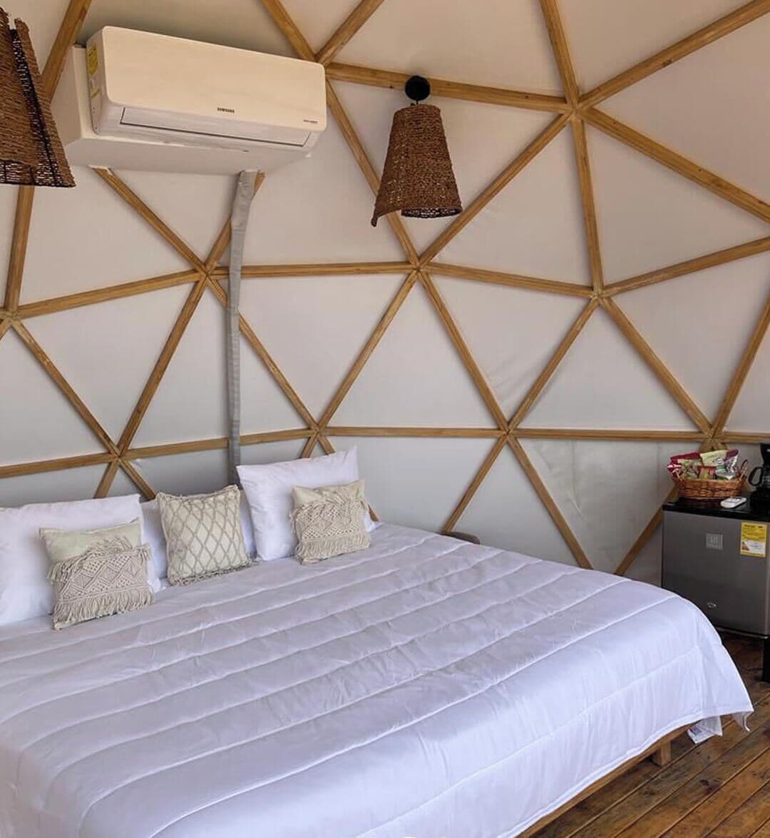 Una cama con una colcha blanca y varios almohadones, una pared con diseño geométrico de madera, y un ventilador de techo.