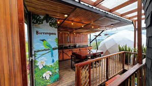 Un área de comedor al aire libre con una terraza de madera, un mural con aves y hojas tropicales, y vistas al paisaje.
