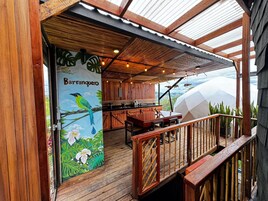 Un área de comedor al aire libre con una terraza de madera, un mural con aves y hojas tropicales, y vistas al paisaje.