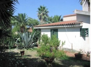 Ein kleines Gebäude mit rotem Ziegeldach, umgeben von viel Grün, darunter Palmen und eine große Agave.