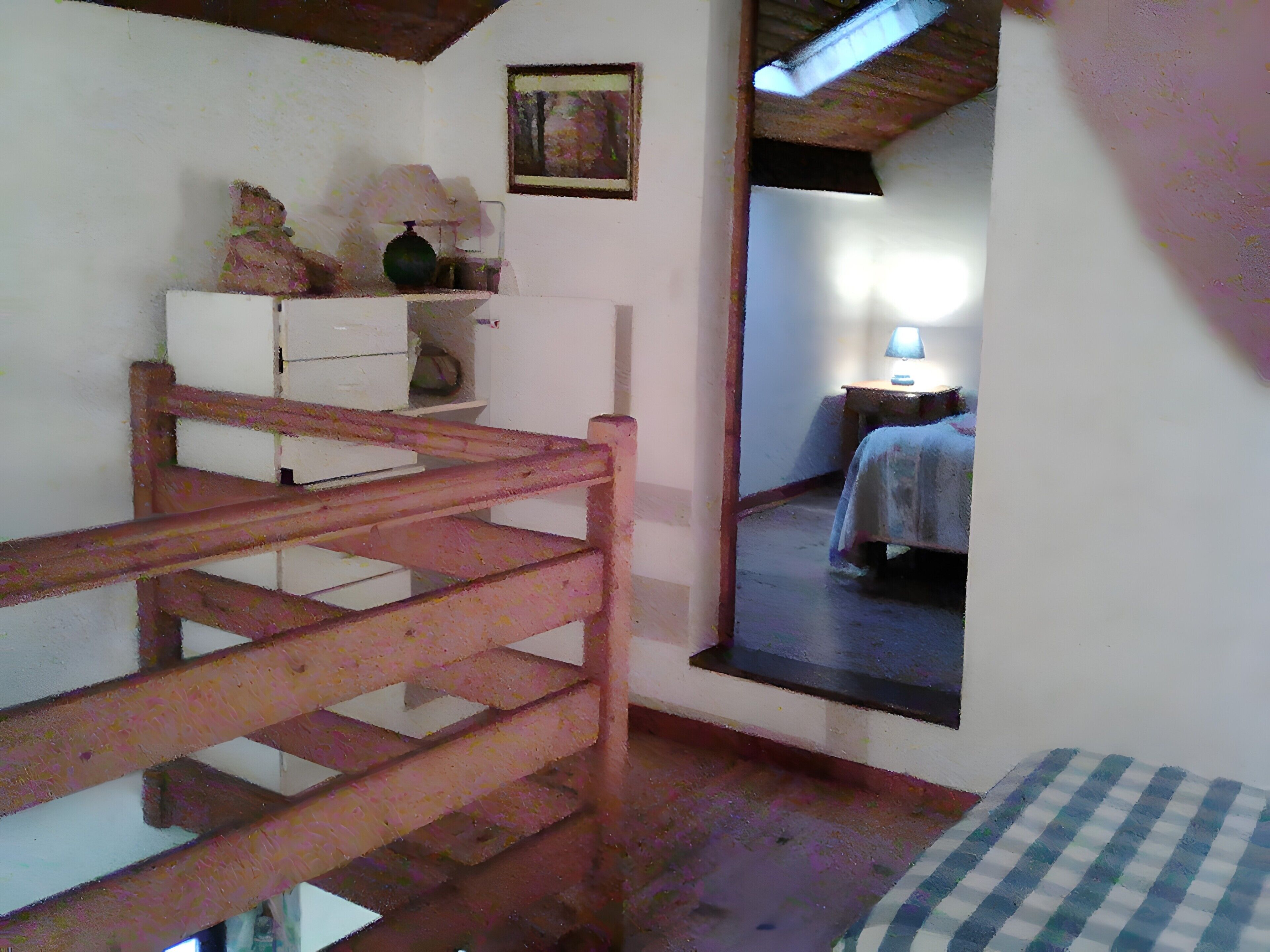A room with a wooden staircase, a bed with a checkered bedspread, a nightstand with a lamp, and a painting on the wall.