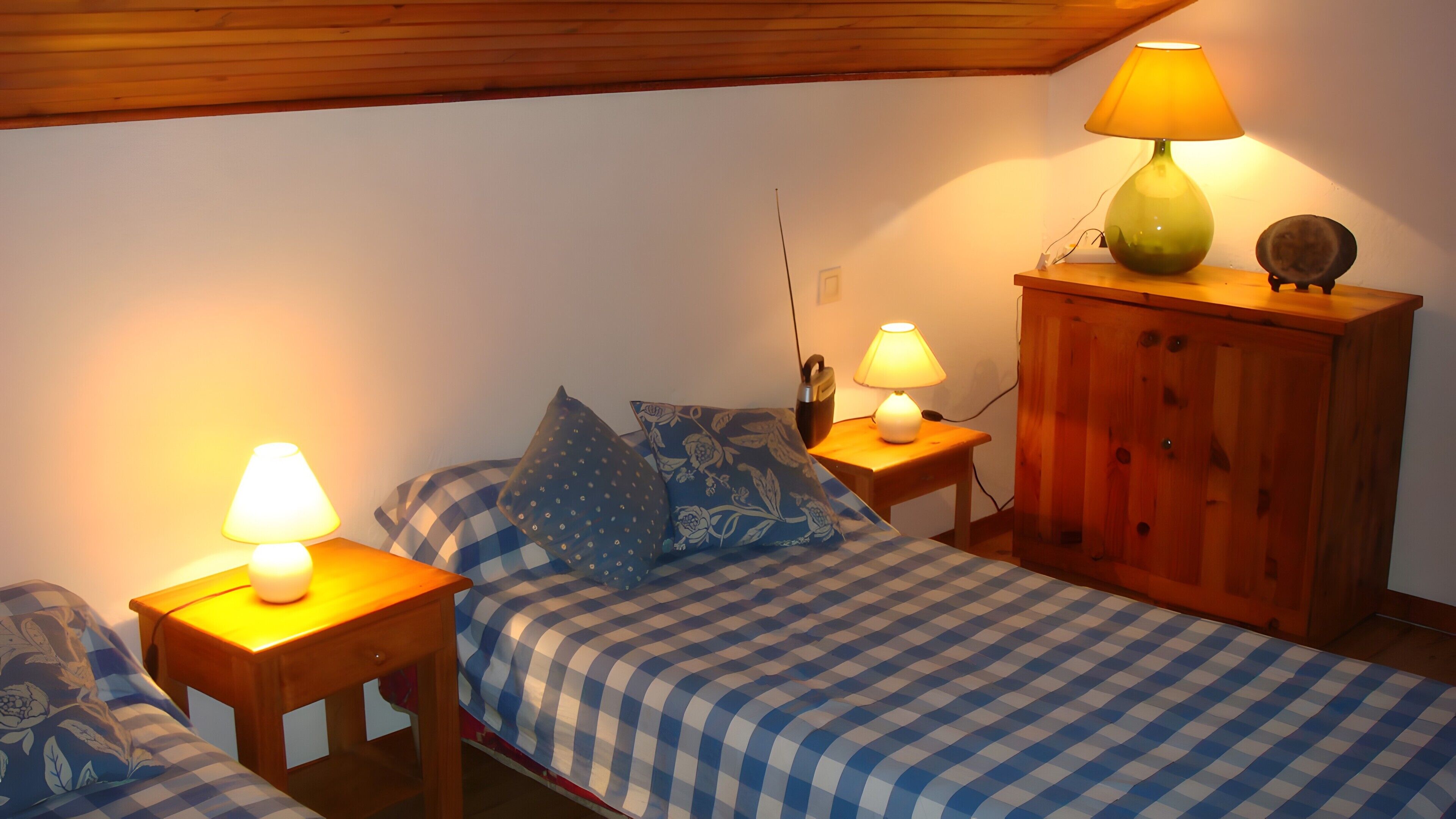 Une chambre à coucher avec une couvre-lit à carreaux, deux tables de chevet avec des lampes, une armoire en bois et une lampe fixée au mur.
