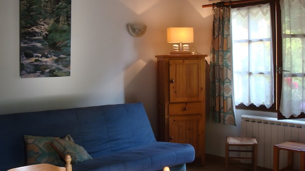 A room with a blue sofa, a wooden cabinet with a lamp, a small stool, and a window with patterned curtains. - Holiday Home 'Gite Des Carcis' with Mountain View, Private Terrace and Wi-Fi (Ganac)
