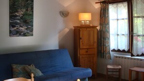 A room with a blue sofa, a wooden cabinet with a lamp, a small stool, and a window with patterned curtains.