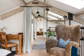 Ein Loft-Zimmer mit hohen Decken, freiliegenden Balken, einem gemütlichen Sessel mit gemustertem Kissen, einem Holztisch mit Stuhl und einem Bett mit weißem Kopfteil.