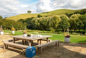 An outdoor dining area with a wooden table and benches, surrounded by a landscaped garden with a hill in the background.