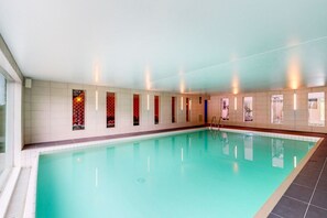 A large indoor swimming pool with a tiled floor and walls, surrounded by windows and a ceiling with recessed lighting.