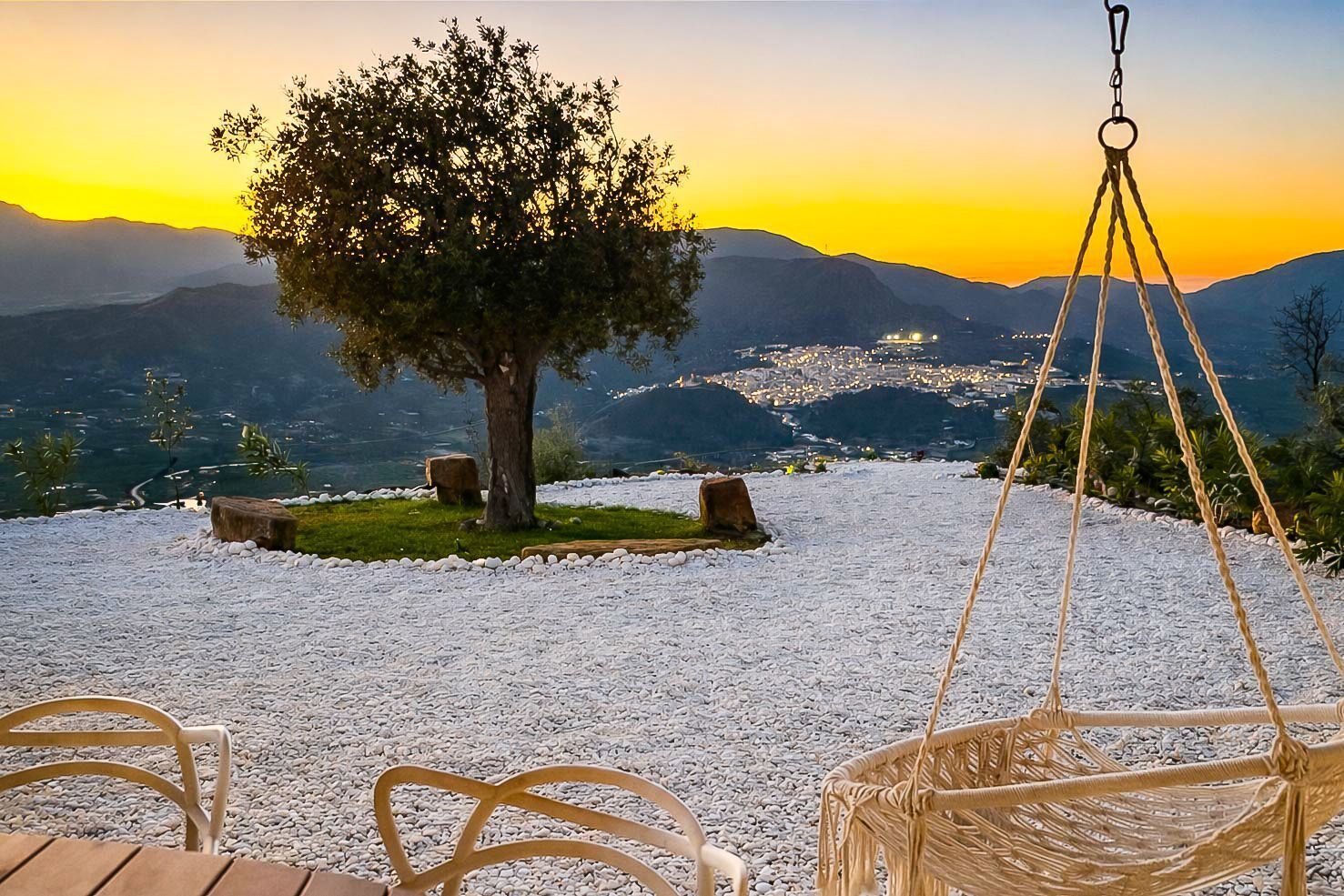 A serene outdoor seating area with a tree, a hanging chair, and a view of a cityscape at sunset.