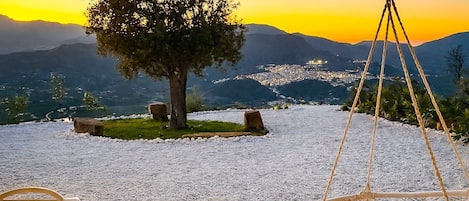 Ein ruhiger Sitzbereich im Freien mit einem Baum, einem Hängestuhl und Blick auf eine Stadtlandschaft bei Sonnenuntergang.