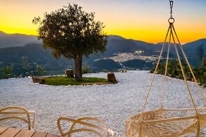 A serene outdoor seating area with a tree, a hanging chair, and a view of a cityscape at sunset. - Villa Alora Lux - Two Bedroom Villa, Sleeps 6 (Álora)