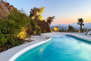 Piscina com borda curva, rodeada por pedras e plantas, exibindo vista para montanhas distantes e um pôr do sol.