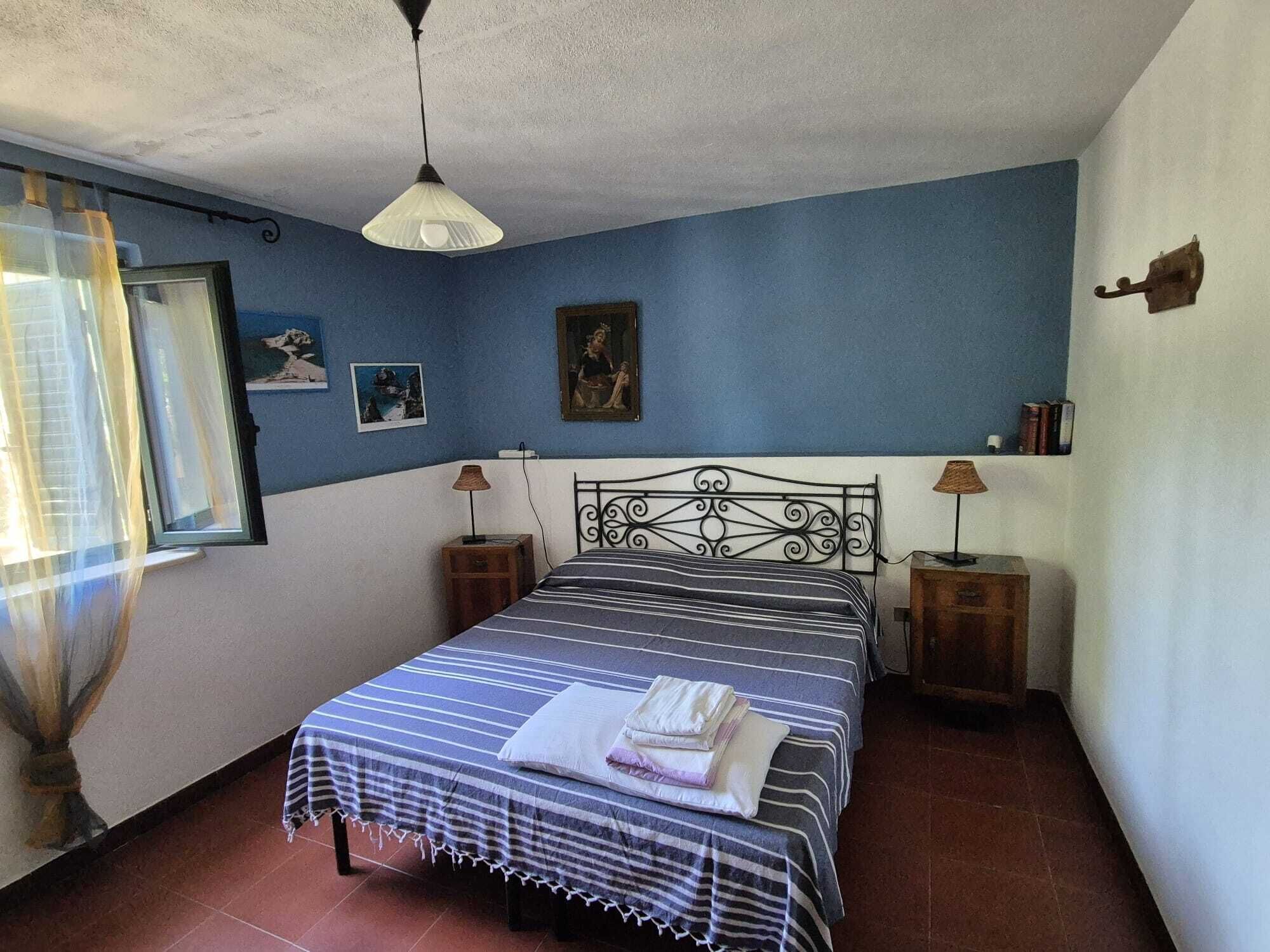 A bedroom with a bed, two bedside tables, a hanging light, a window with curtains, and wall art.