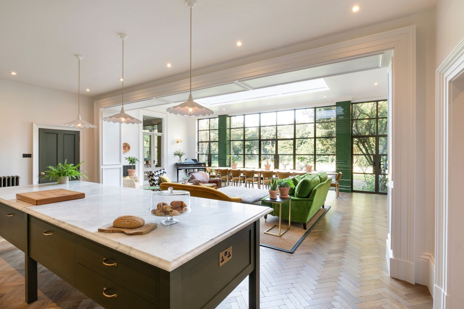 A spacious kitchen with a large island, dining area with green sofas, and a view through large windows.