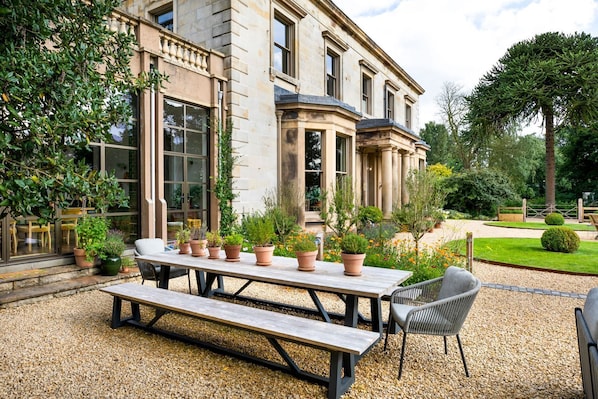 A stone building with a patio area featuring a wooden table and chairs, surrounded by potted plants and a well-maintained garden.