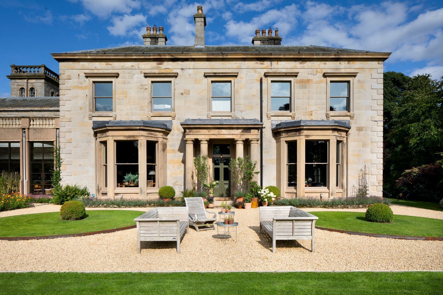 A large stone building with a symmetrical facade, large windows, and a well-maintained garden with a gravel path and two lounge chairs.