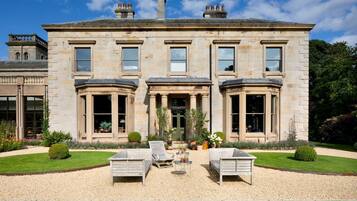 A large stone building with a symmetrical facade, large windows, and a well-maintained garden with a gravel path and two lounge chairs.