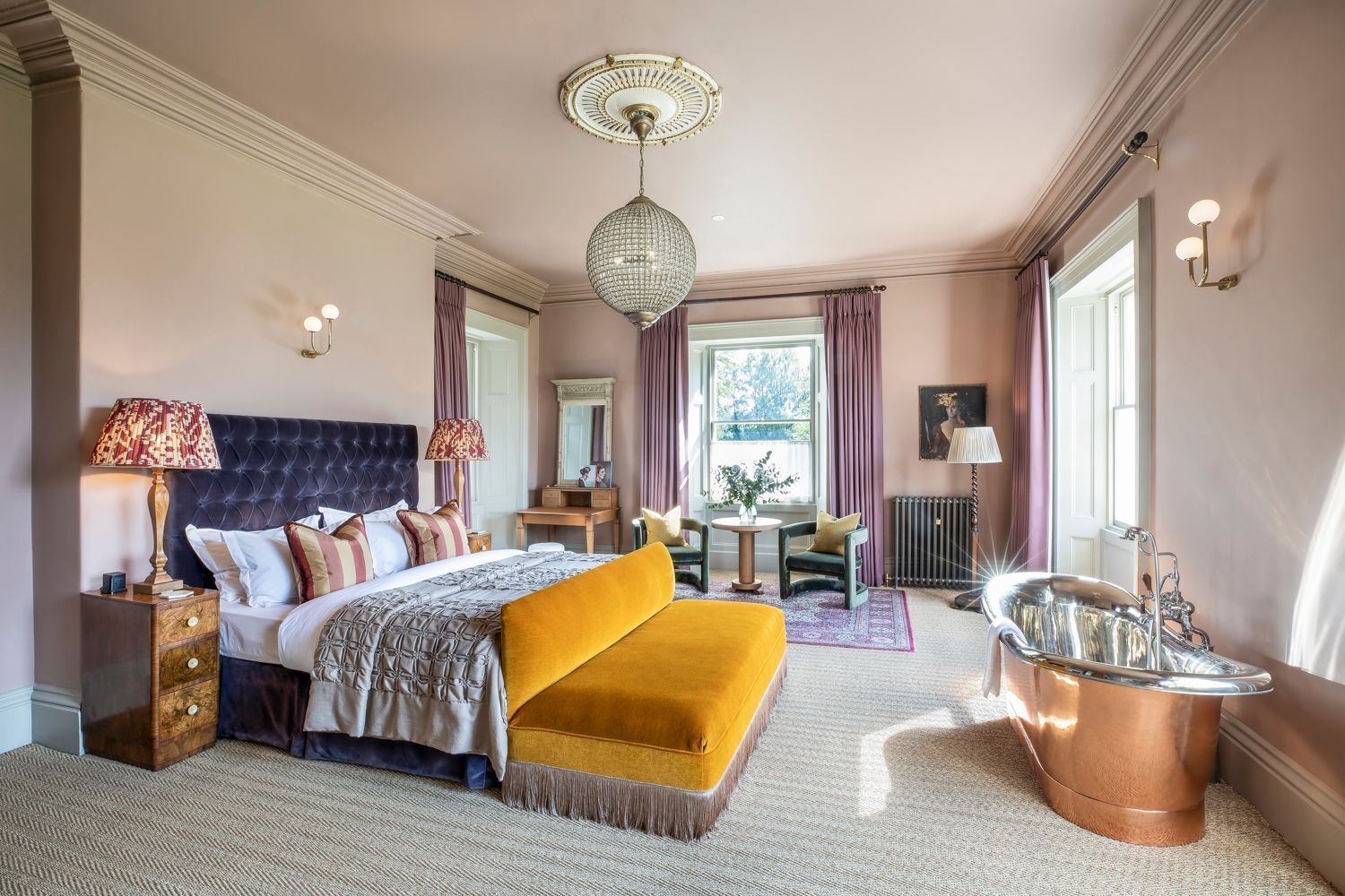 A bedroom with a large bed, a sitting area, a vanity with a mirror, and a bathtub.