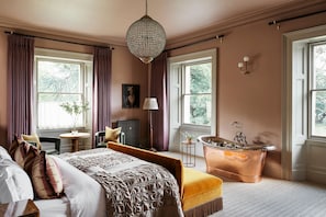 A bedroom with a large bed, a sitting area with a chair and a small table, a bathtub, and large windows with curtains.