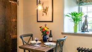 A dining table with chairs, a framed botanical print, a vase with flowers, a bottle of wine, and a window with a view of greenery.
