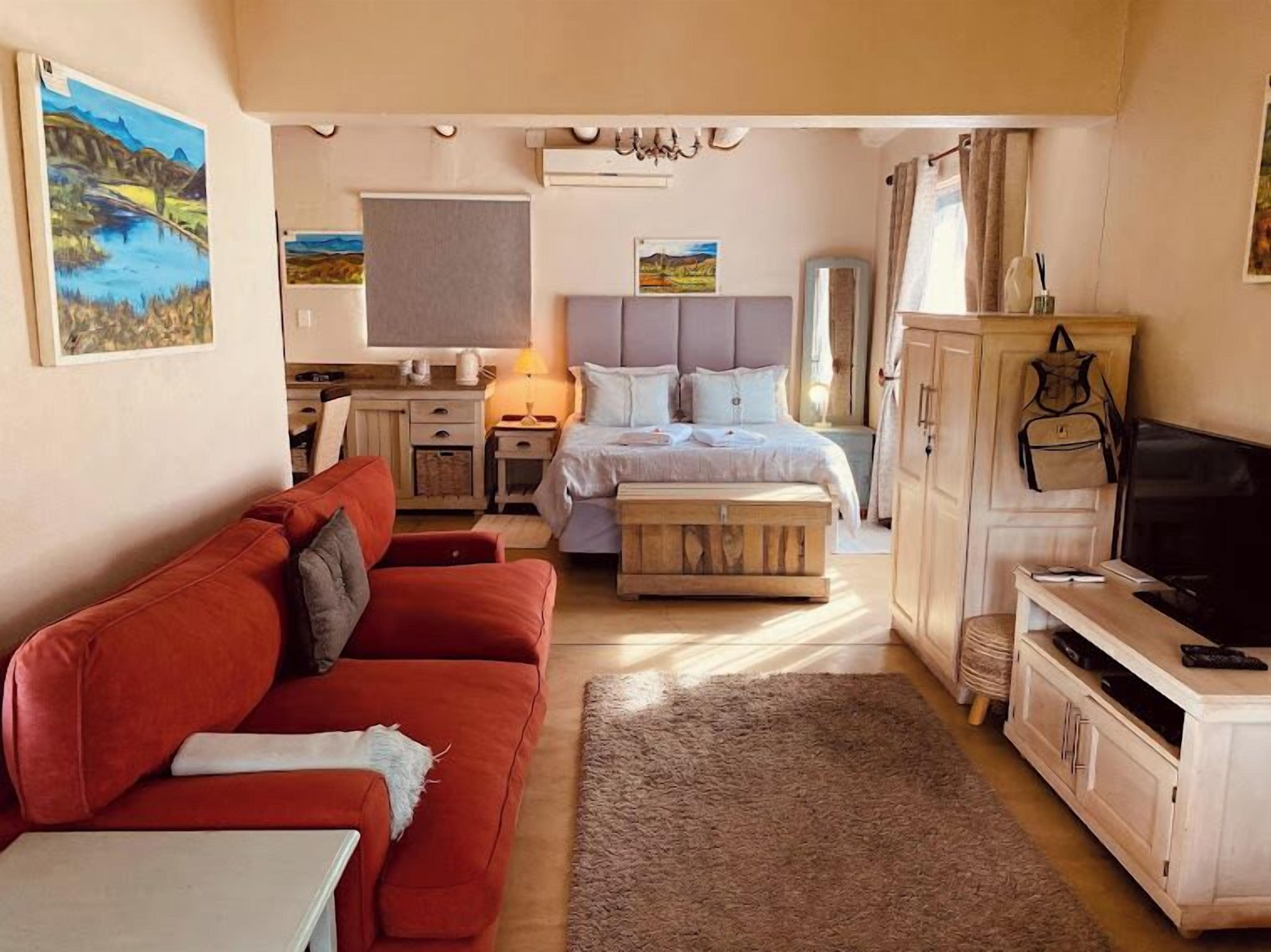 Una habitación compacta con un sofá rojo, una cama con cabecera gris, un televisor y una mesita.
