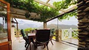 A covered patio with a table and chairs, a stone wall, and a view of mountains. - Oaksrest Vineyards Guest Farm (Ladismith)