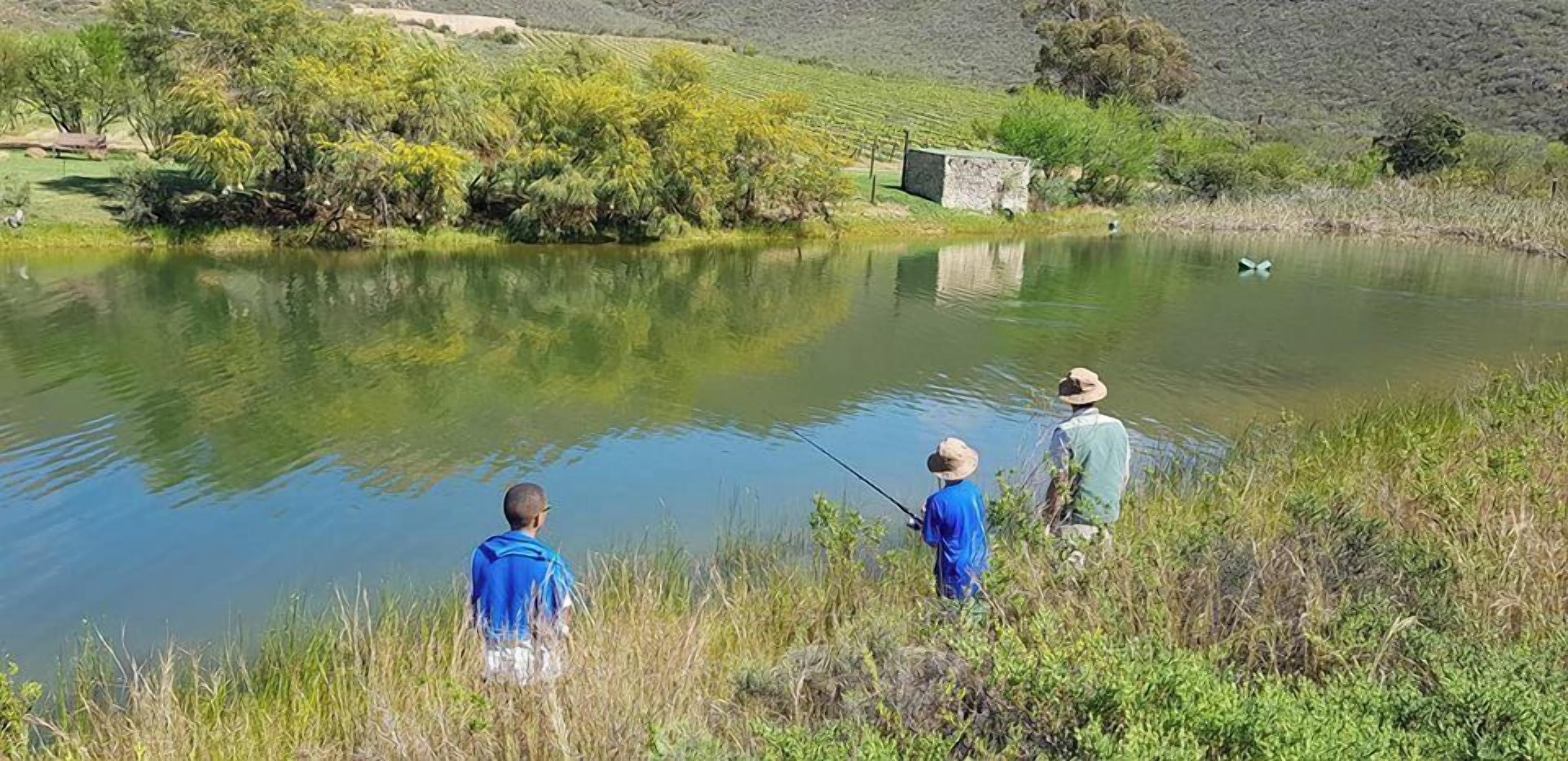 A calm lake with people fishing, surrounded by greenery and a stone structure.