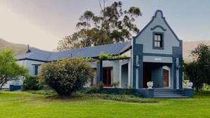 A two-story house with a prominent front porch, a well-maintained lawn, and a mountain backdrop. - Oaksrest Vineyards Guest Farm (Ladismith)