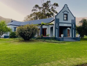 A two-story house with a prominent front porch, a well-maintained lawn, and a mountain backdrop. - Oaksrest Vineyards Guest Farm (Ladismith)