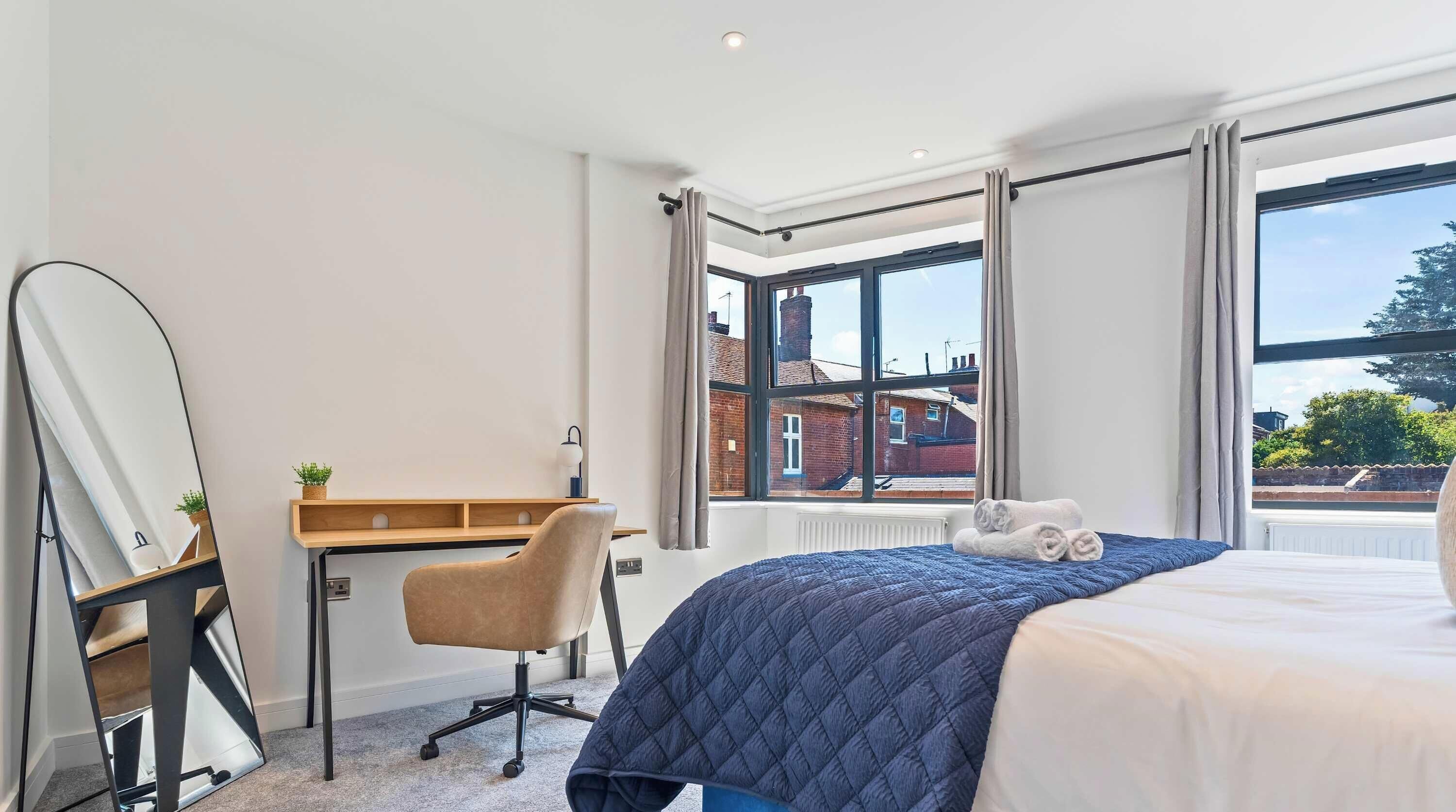 A bedroom with a bed, a desk, and a chair. There is a window with curtains, and a view of buildings outside.
