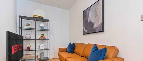 A living room with a brown sofa, a glass coffee table, a TV on a stand, a bookshelf with various items, and a framed picture on the wall.