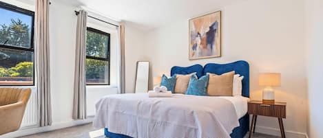 A hotel room with a large bed, a side table with a lamp, a chair, and a view of greenery through the windows.